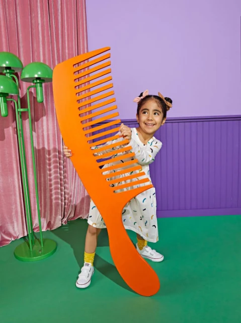 Young girl smiles while holding a large orange comb, standing indoors with a purple wall and green floor, adding a whimsical touch.