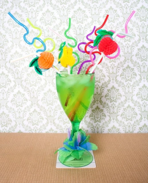 Vibrant green cocktail in a glass with colorful straws and fruit decorations, set against a patterned wallpaper backdrop.
