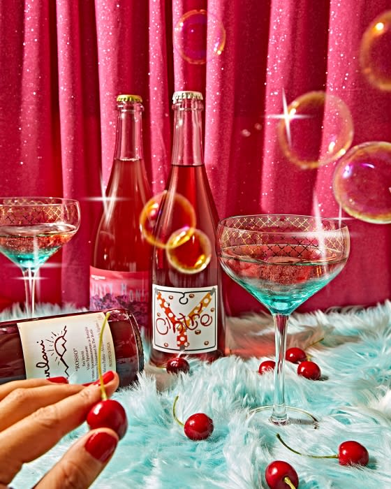 Two wine bottles and stemmed glasses with cherries are displayed on a furry surface in front of a red curtain backdrop.