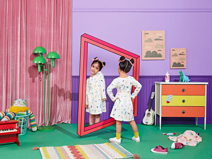 Young girl in a patterned dress poses in front of a rectangular mirror in a brightly colored playroom, with a drawn pink frame.