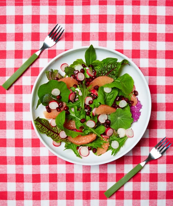 Overhead shot of a fresh salad with grapefruit, radish, and greens on a white plate, with two forks on a red and white tablecloth.