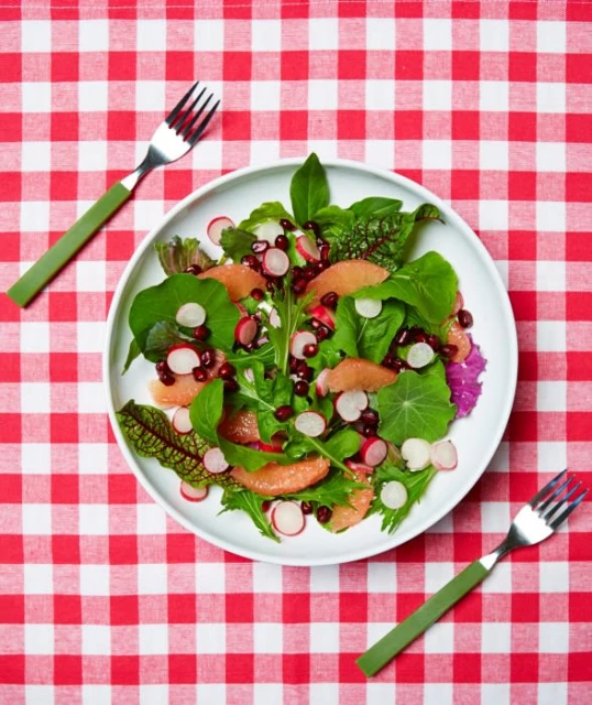 Overhead shot of a fresh salad with grapefruit, radish, and greens on a white plate, with two forks on a red and white tablecloth.