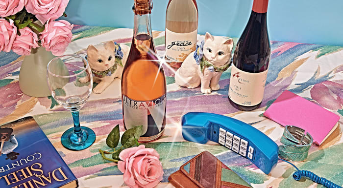 Still life arrangement featuring wine bottles, vintage rotary phone, faux cat figurines, books, and pink flowers on a floral tablecloth.