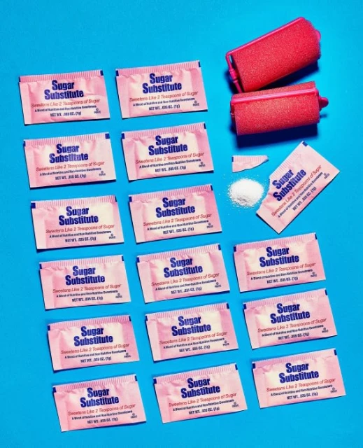 Arrangement of sugar substitute packets and pink hair rollers on a bright blue background, creating a vibrant and contrasting aesthetic.