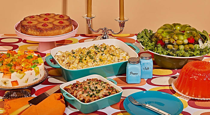 Retro dinner table with cake, casseroles, jello salad, and jello molds arranged on a patterned tablecloth in a studio setting.