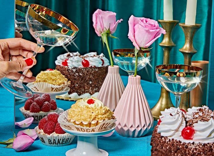 Festive still life featuring an array of cakes, pink roses in vases, and elegant glasses on a teal tablecloth against a teal curtain.