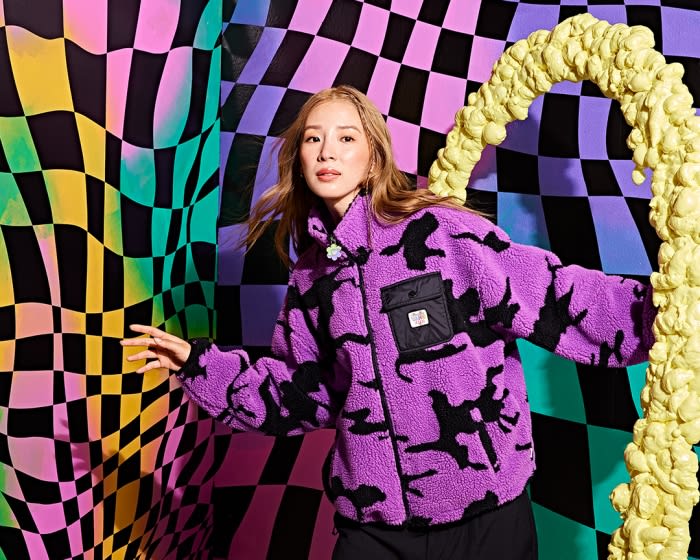 Woman in a purple camouflage jacket poses in front of a multicolored checkered wall with a yellow foam prop, creating a vibrant studio portrait.