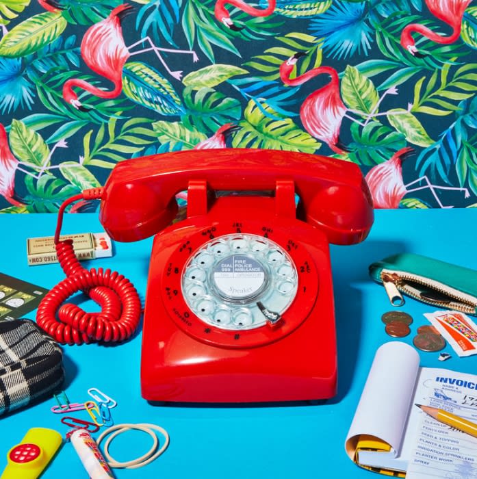 Retro red rotary phone with coiled cord sits on a blue surface amidst scattered office supplies and a vibrant flamingo wallpaper backdrop.