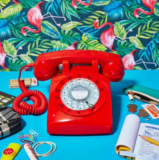 Retro red rotary phone with coiled cord sits on a blue surface amidst scattered office supplies and a vibrant flamingo wallpaper backdrop.