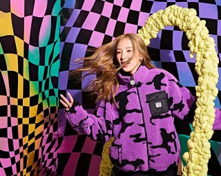 Young woman with blonde hair wearing a purple and black fleece jacket in front of a distorted checkerboard backdrop.