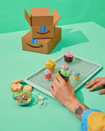 Person holding decorated cupcake near cardboard shipping boxes and baking supplies on a green surface