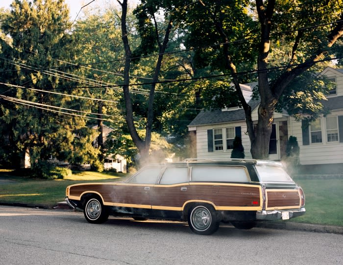 Classic wood-paneled station wagon parked on a residential street, with green trees and a house in the background under daylight.