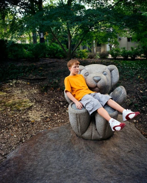 Boy sitting on a stone bear sculpture in a park setting with green trees and foliage in the background, wearing shorts and orange t-shirt.