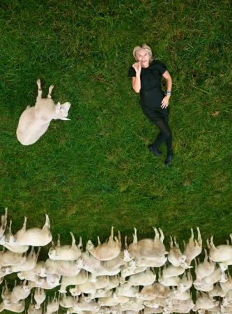 Overhead portrait of a blonde woman lying on green grass near a large flock of white sheep by photographer Chris Buck