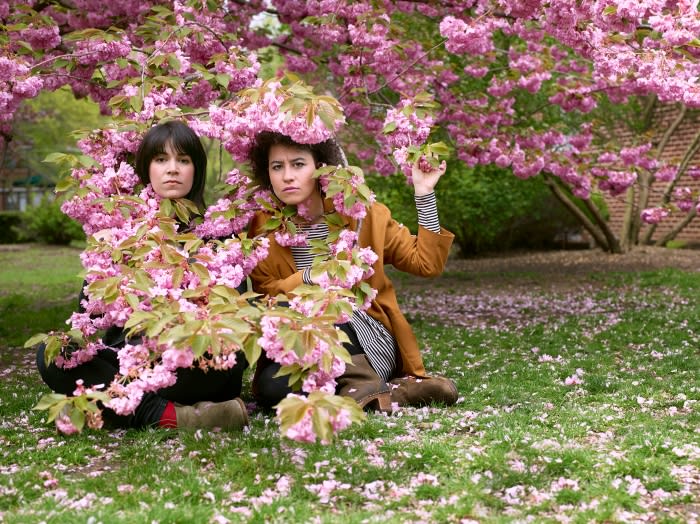 Two women sit on grass surrounded by pink blossoms from cherry trees in full bloom, creating a natural springtime portrait.