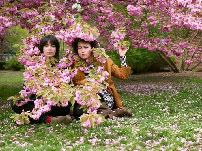 Two women sit on grass surrounded by pink blossoms from cherry trees in full bloom, creating a natural springtime portrait.