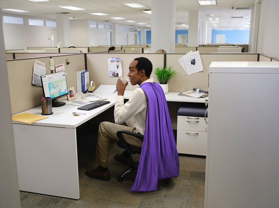 Man in purple cape sitting at office desk with computer, drinking from cup, in cubicle setting with beige and white decor.