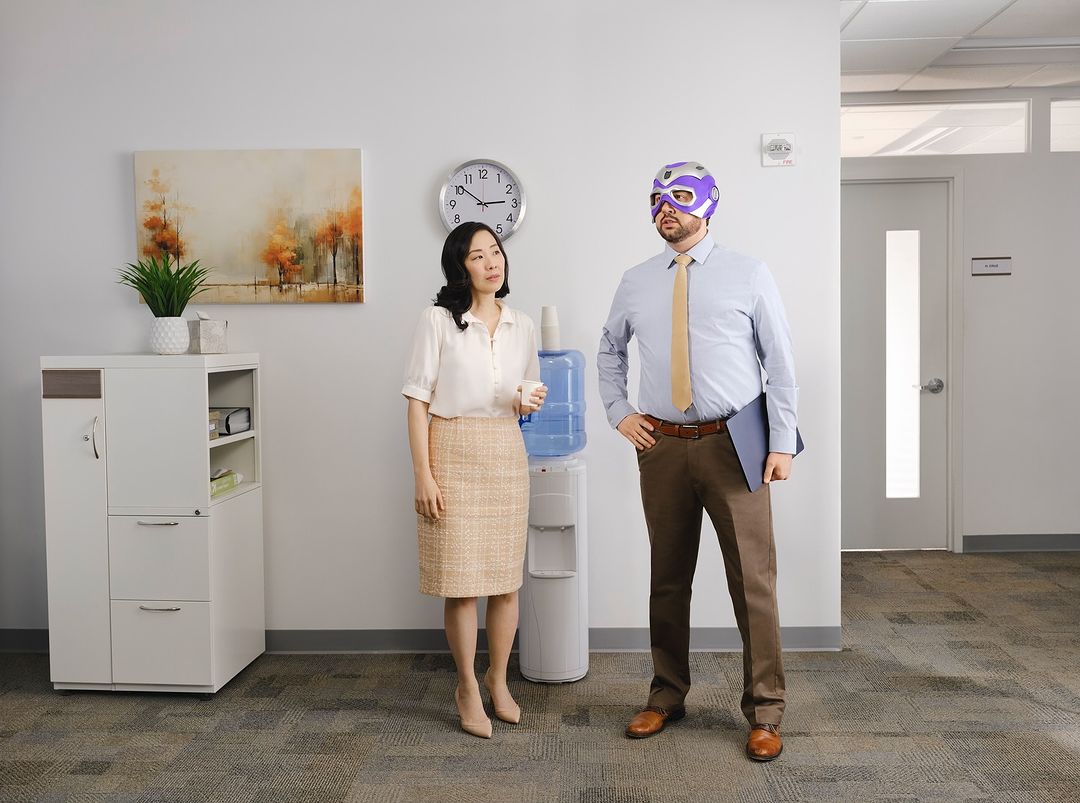 An Asian woman stands near a water cooler in an office, while a Caucasian man with a purple mask stands ready to defend against thirst.