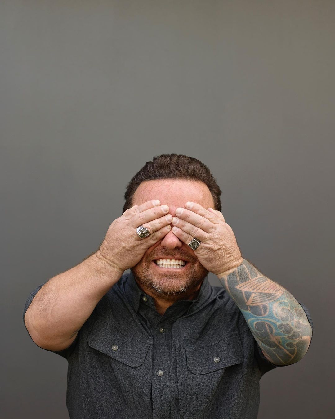 An adult man covers his eyes with both hands in a medium studio shot against a gray background, wearing a gray button-down shirt.