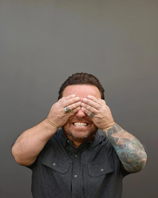 An adult man covers his eyes with both hands in a medium studio shot against a gray background, wearing a gray button-down shirt.