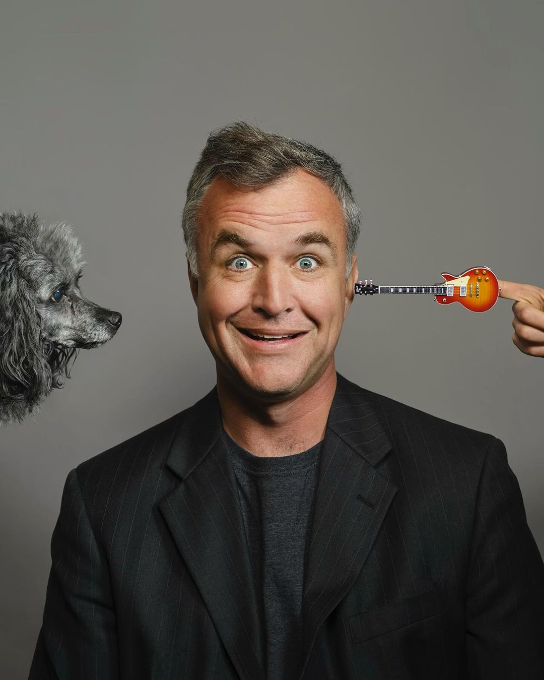 Portrait of a smiling man with a toy guitar next to his head, and a gray poodle on the left side against a grey background.
