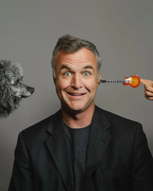 Portrait of a smiling man with a toy guitar next to his head, and a gray poodle on the left side against a grey background.