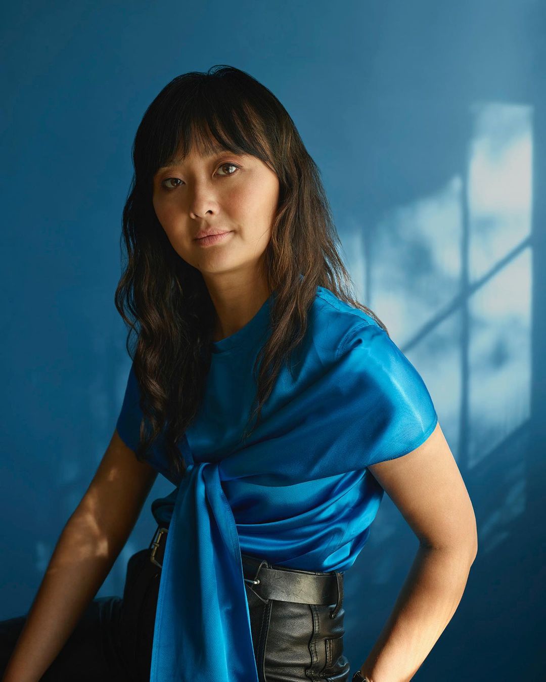 Portrait of a woman with dark hair wearing a blue satin top and black leather pants, posing against a solid blue backdrop with shadows.
