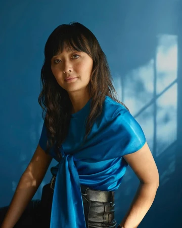 Portrait of a woman with dark hair wearing a blue satin top and black leather pants, posing against a solid blue backdrop with shadows.