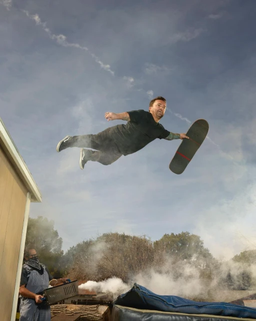Man in mid-air on skateboard with background smoke effect and another man with blower; outdoor skateboarding action shot.