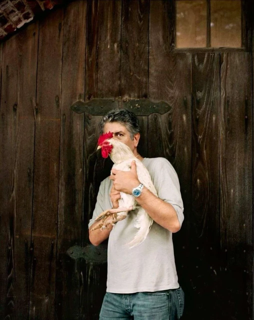 Adult male embraces white rooster in front of rustic wood door. The man wears a grey shirt and jeans; daylight.