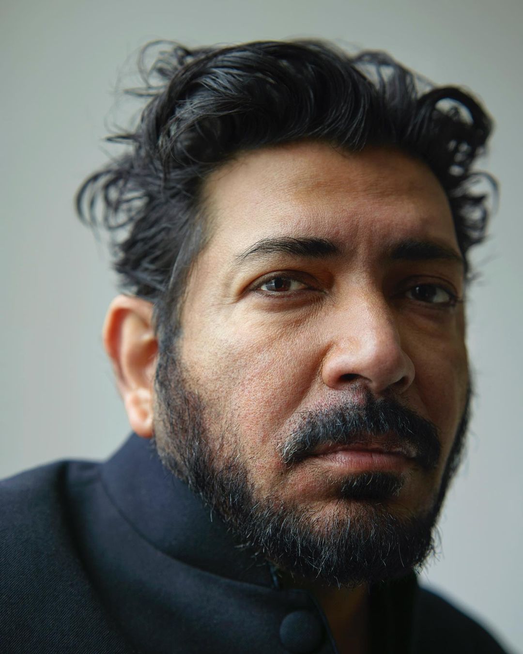 Close-up portrait of a man with dark curly hair and a beard wearing a dark mandarin collar shirt against a plain light wall.