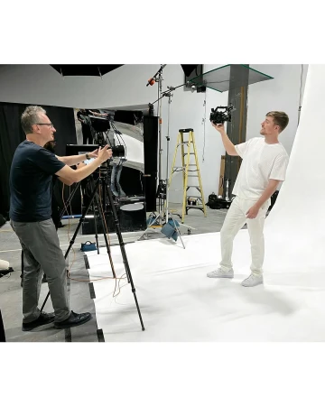 Two adult males in a photo studio. One man holds a camera up while the other adjusts a professional video camera on a tripod.