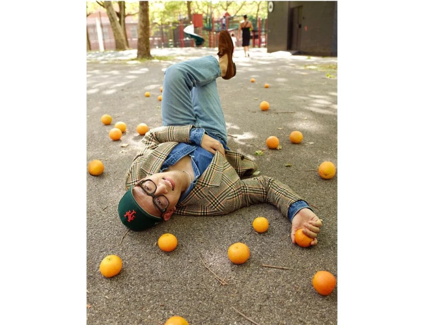 Adult male in a denim shirt, plaid blazer, and cap lies upside down on concrete surrounded by scattered oranges in outdoor setting.