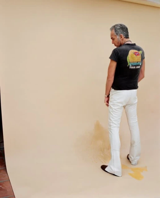 Full shot of adult man in black t-shirt and white pants standing in front of tan backdrop in a photo studio with wet spot at feet.