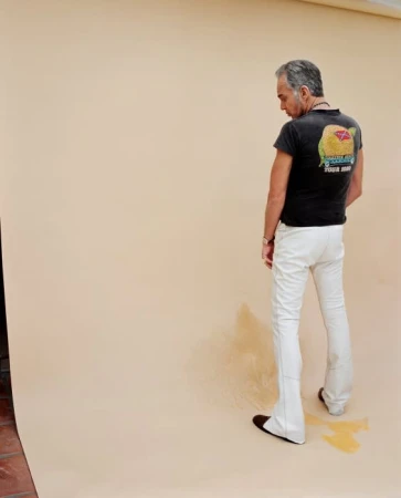 Full shot of adult man in black t-shirt and white pants standing in front of tan backdrop in a photo studio with wet spot at feet.