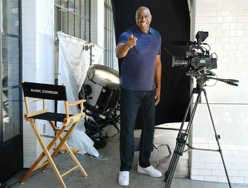 An adult Black man points at the camera in a light-filled studio, alongside a large camera, lighting, and a director's chair.