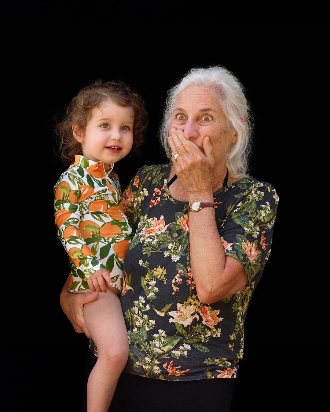 Grandmother holding her granddaughter who is wearing a swimsuit with oranges, looking surprised against a dark background.