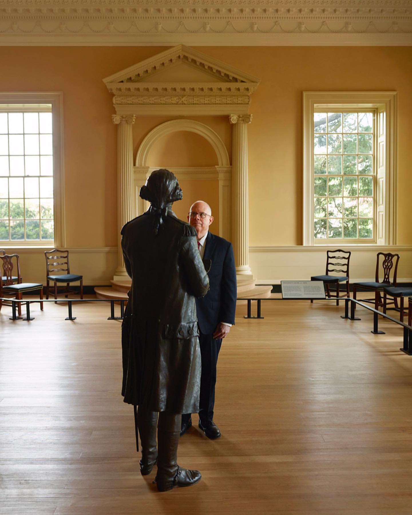 Indoor shot features a bronze statue of a man facing away from the viewer, with a businessman standing nearby in a large hall.