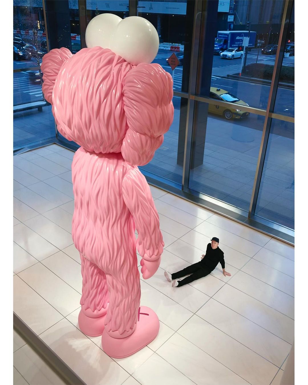 Large pink KAWS sculpture in a modern building with a man lying on the floor, looking up at the art piece through a glass wall.