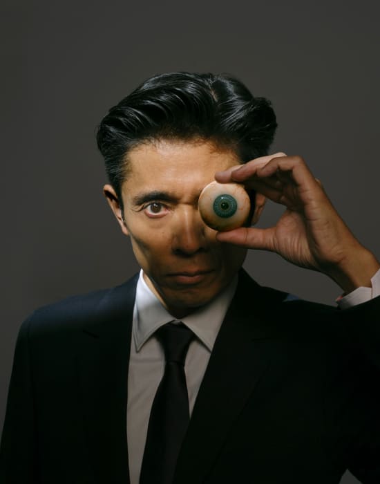Man in suit poses indoors, covering one eye with artificial eyeball.  Formal attire and focused expression are key visual elements.