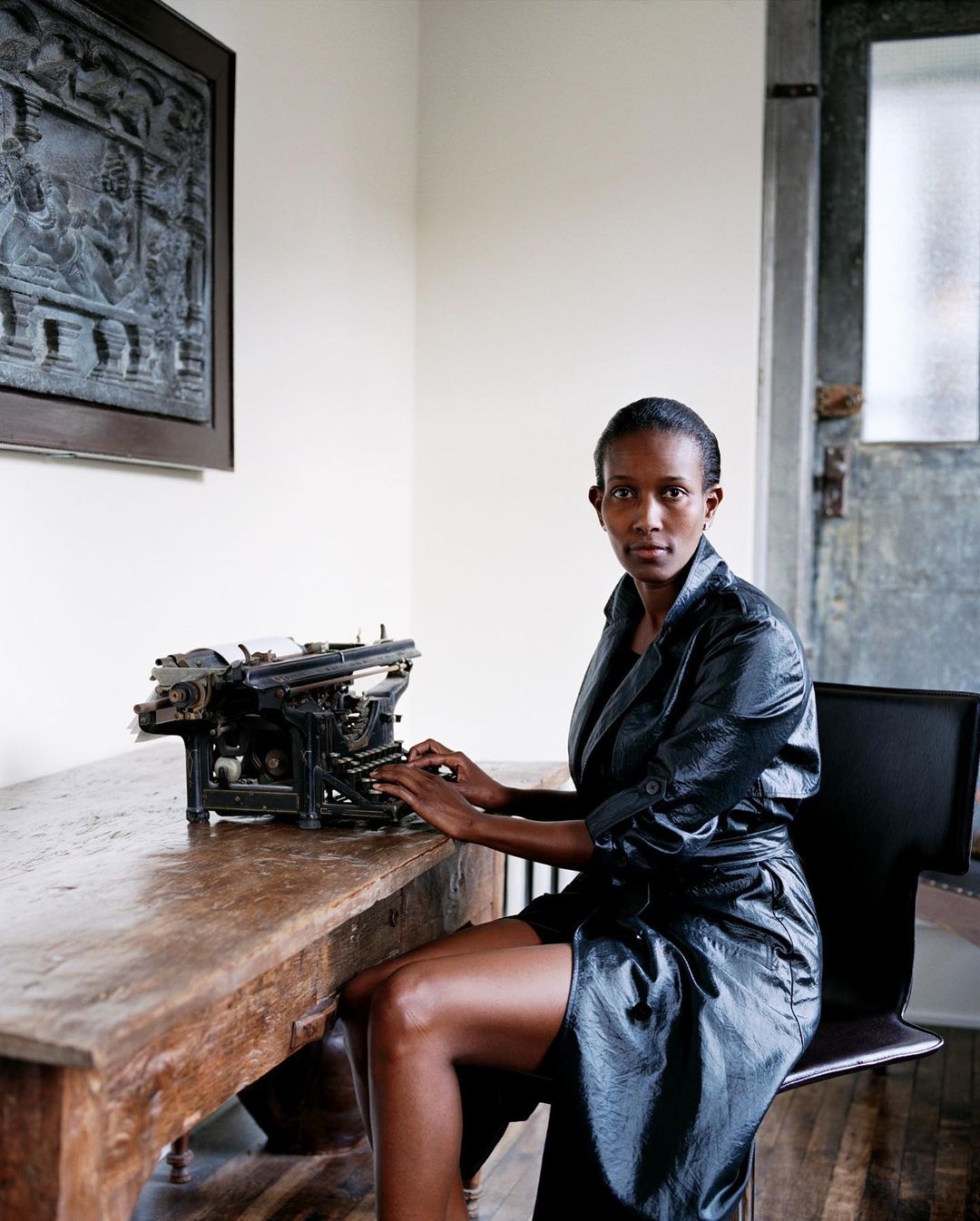 Woman in a black robe sits at a rustic wooden desk using a vintage typewriter in a minimalist indoor setting with natural light.
