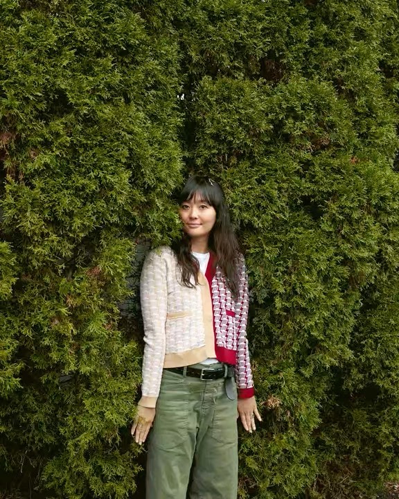 An Asian woman with dark hair stands in front of green shrubbery, wearing a mismatched cardigan and olive green pants in an outdoor setting.