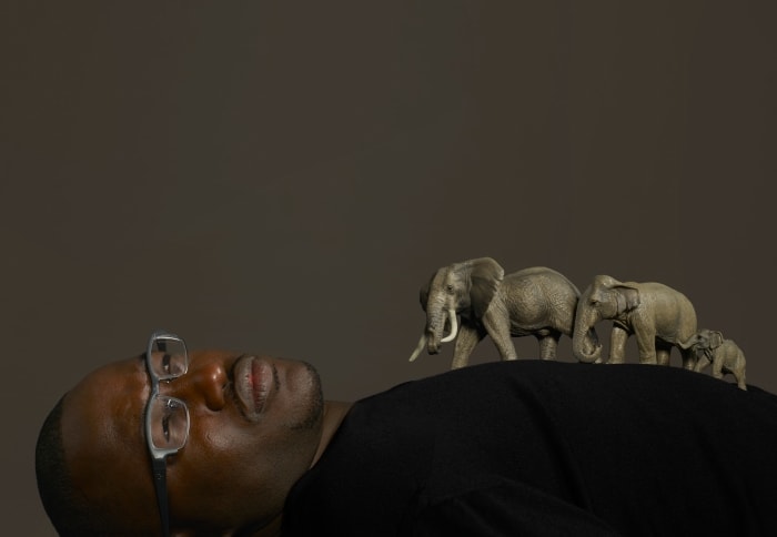 Portrait of an African American man lying down with glasses, supporting a line of toy elephants on his body in a studio setting.