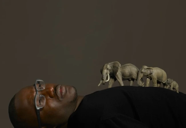 Portrait of an African American man lying down with glasses, supporting a line of toy elephants on his body in a studio setting.
