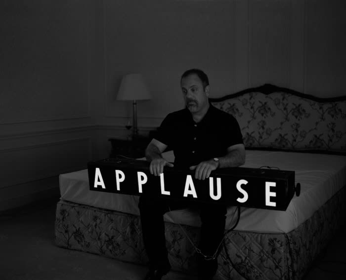Monochrome portrait of a man sitting in a bedroom, holding a light-up sign reading 'APPLAUSE'. Lamp and bed are in the background.