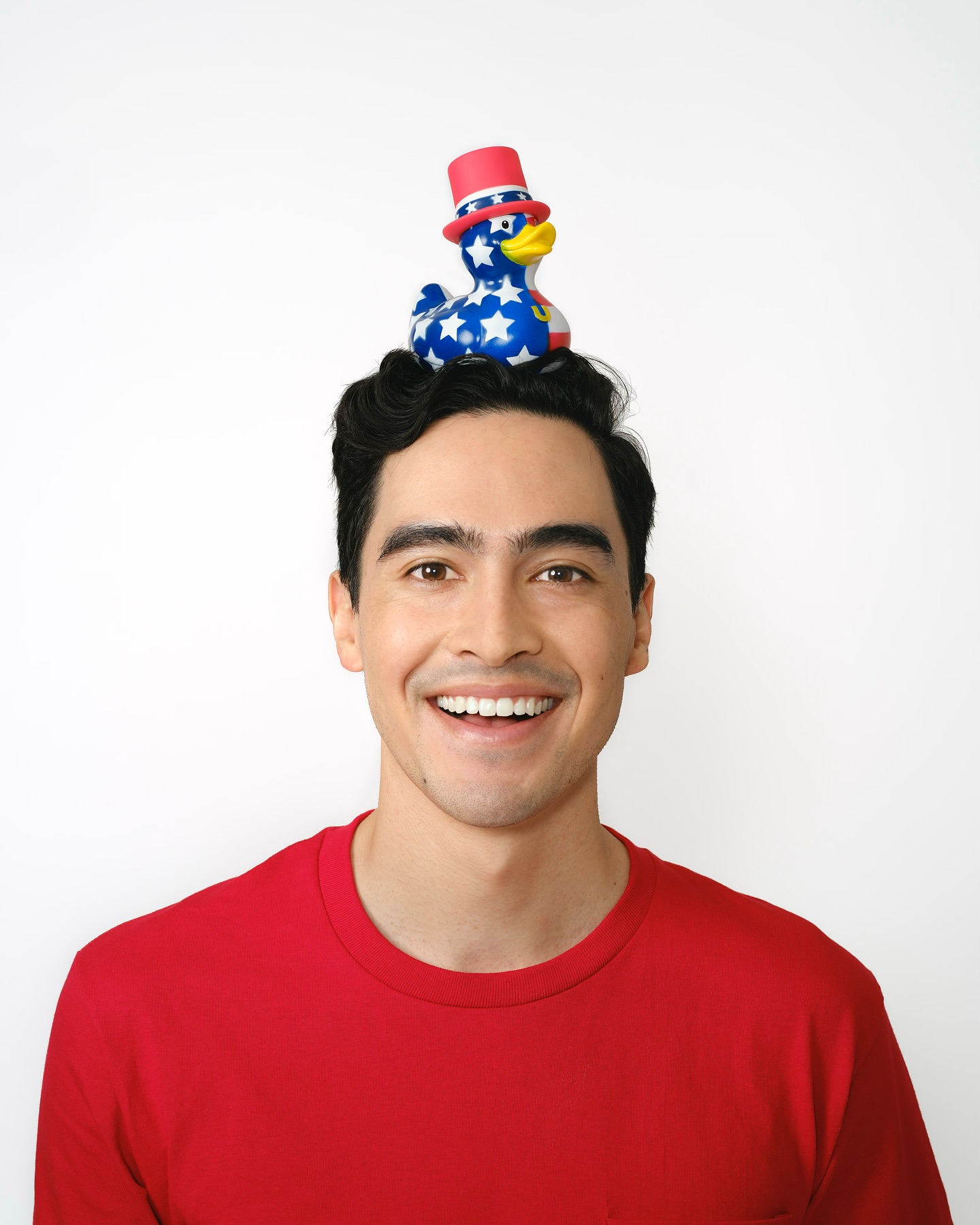 Smiling man in red shirt poses with patriotic rubber duck wearing top hat balanced on head against white background in eye-level medium shot.
