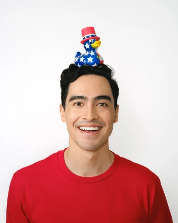 Smiling man in red shirt poses with patriotic rubber duck wearing top hat balanced on head against white background in eye-level medium shot.
