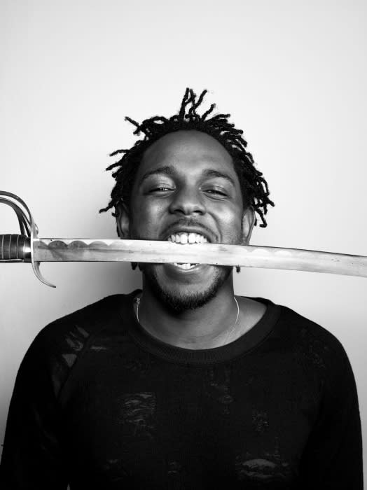 Monochrome studio portrait of a smiling Black man with dreadlocks holding a sword in his mouth against a white backdrop.