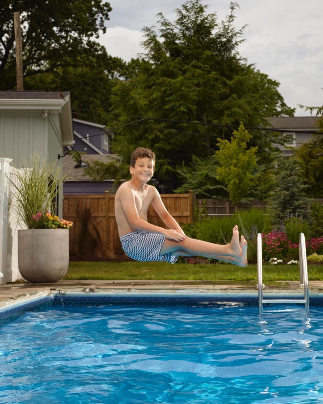 A young boy leaps joyfully over a shimmering blue swimming pool in a backyard setting, capturing a moment of summer fun and recreation.