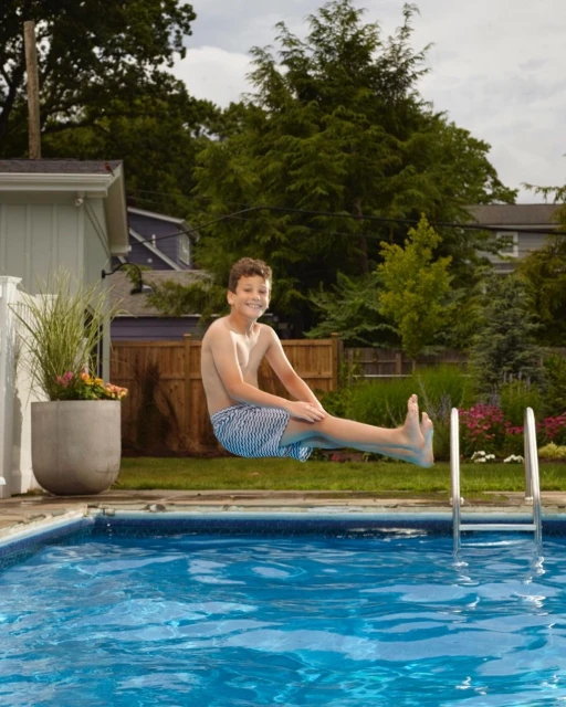 A young boy leaps joyfully over a shimmering blue swimming pool in a backyard setting, capturing a moment of summer fun and recreation.